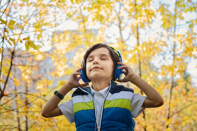 Chłopiec stojący na tle jesiennych drzew i słuchający muzyki przez niebieskie, bezprzewodowe słuchawki nauszne.