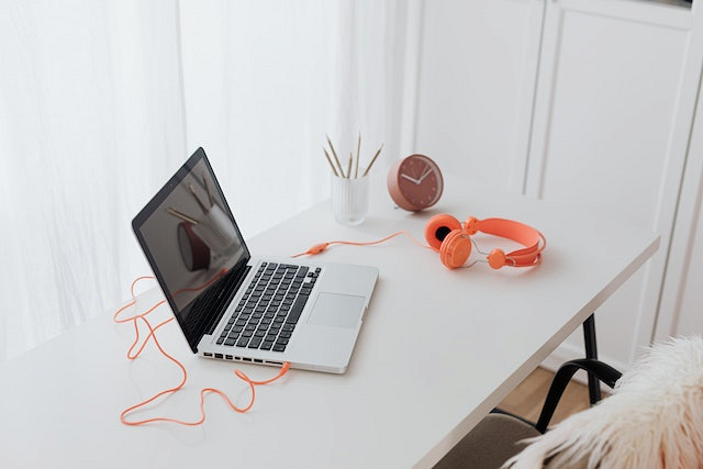 Pomarańczowe, przewodowe słuchawki nauszne leżą na białym blacie biurka, podłączone do otwartego laptopa.